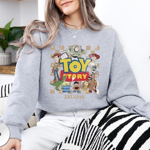 May include: A grey crewneck sweatshirt featuring a graphic print of Toy Story characters including Woody, Buzz Lightyear, Jessie, Rex, Slinky Dog, Mr. Potato Head, and an Alien. The design includes the text TOY STORY and EST. 1995 on a checkered background.