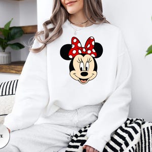Puede incluir: Sudadera blanca con un gr&aacute;fico de Minnie Mouse. El gr&aacute;fico muestra la cara de Minnie con un lazo rojo con lunares blancos. La sudadera es de cuello redondo y est&aacute; hecha de un material suave. La palabra "WHITE" est&aacute; impresa en un c&iacute;rculo blanco.