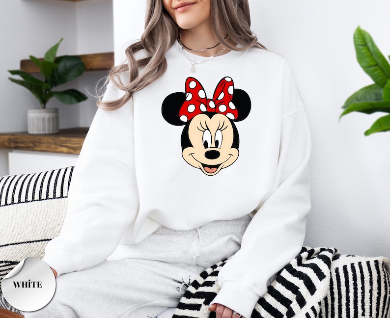 Puede incluir: Sudadera blanca con un gr&aacute;fico de Minnie Mouse. El gr&aacute;fico muestra la cara de Minnie con un lazo rojo con lunares blancos. La sudadera es de cuello redondo y est&aacute; hecha de un material suave. La palabra "WHITE" est&aacute; impresa en un c&iacute;rculo blanco.