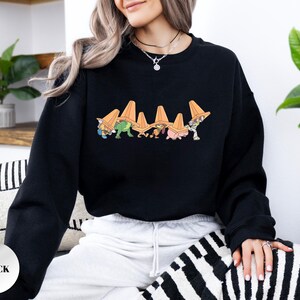 Op de afbeelding: Zwart sweatshirt met een kleurrijke cartoonafbeelding van figuren met oranje verkeerskegels. De afbeelding staat in het midden van de borst. Het sweatshirt heeft een ronde hals.