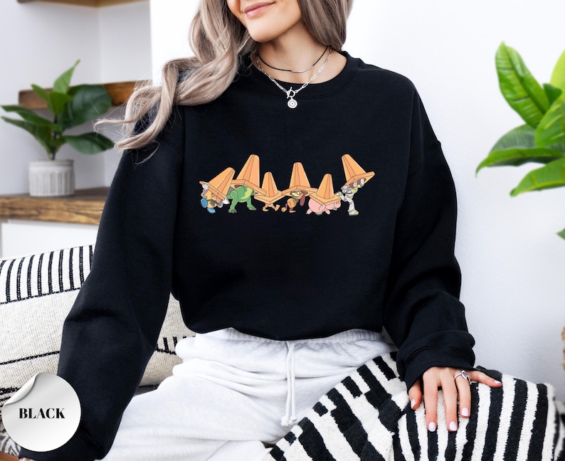 Op de afbeelding: Zwart sweatshirt met een kleurrijke cartoonafbeelding van figuren met oranje verkeerskegels. De afbeelding staat in het midden van de borst. Het sweatshirt heeft een ronde hals.