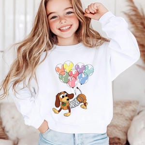 May include: White sweatshirt featuring a cartoon image of Slinky Dog from Toy Story holding colorful Mickey Mouse balloons. The design includes red, yellow, blue, pink, and green balloons. The sweatshirt is a crew neck style.