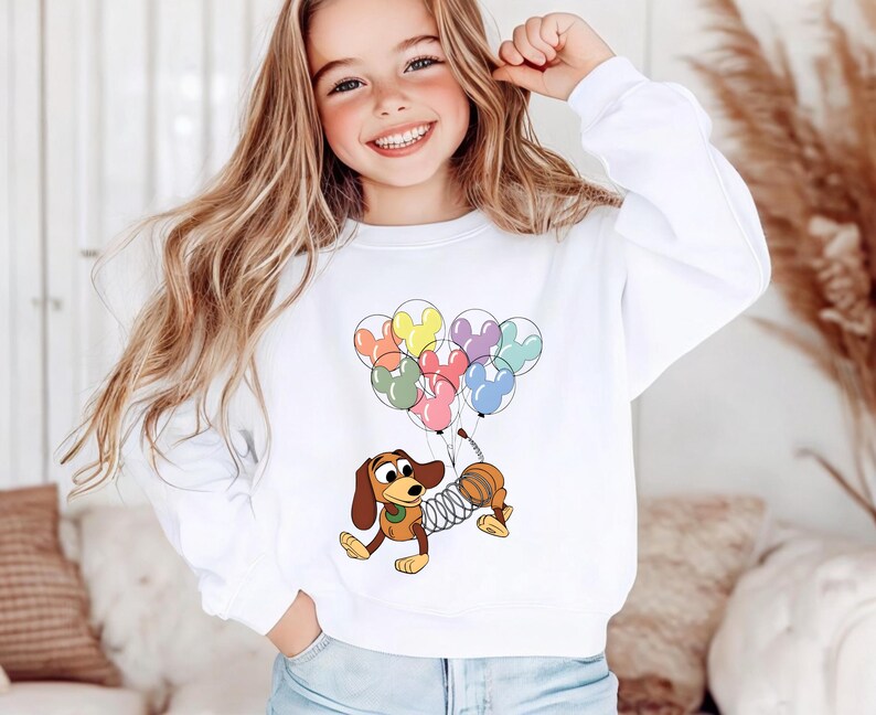 May include: White sweatshirt featuring a cartoon image of Slinky Dog from Toy Story holding colorful Mickey Mouse balloons. The design includes red, yellow, blue, pink, and green balloons. The sweatshirt is a crew neck style.