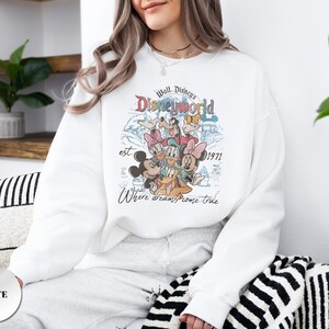 Op de afbeelding: Witte sweatshirt met een kleurrijke afbeelding van Disney-personages, waaronder Mickey en Minnie Mouse, Donald Duck en Goofy. De afbeelding bevat de tekst "Walt Disney's Disneyworld est. 1971" en "Where dreams come true."