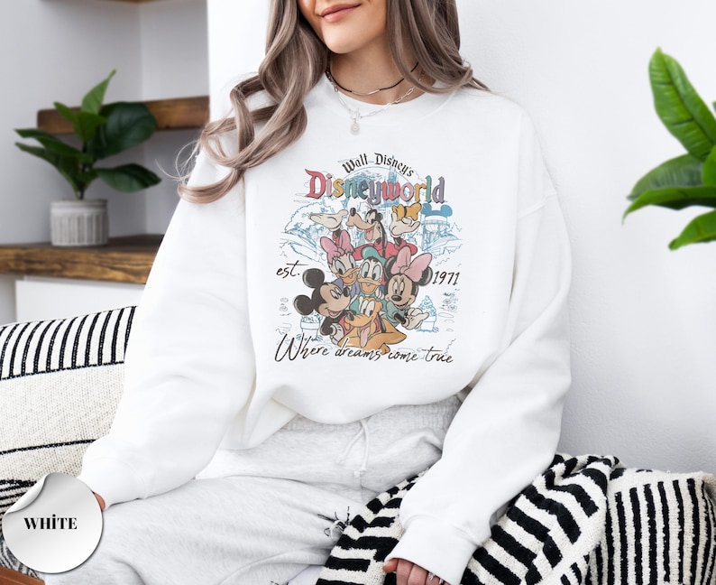 Op de afbeelding: Witte sweatshirt met een kleurrijke afbeelding van Disney-personages, waaronder Mickey en Minnie Mouse, Donald Duck en Goofy. De afbeelding bevat de tekst "Walt Disney's Disneyworld est. 1971" en "Where dreams come true."