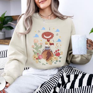 K&ouml;nnte beinhalten: Ein sandfarbenes Sweatshirt mit Rundhalsausschnitt und einem Grafikdruck einer rothaarigen Cowgirl-Figur mit Hut auf einem Pferd, umgeben von Kakteen, Blumen und Noten. Das Design zeigt blaue Wolken und gelbe Sterne auf dem hellbeigen Stoff.