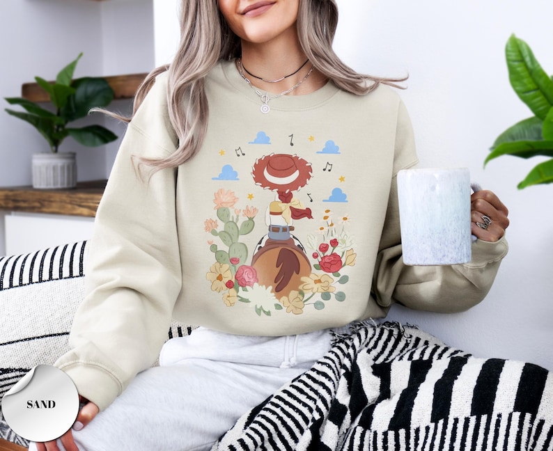 K&ouml;nnte beinhalten: Ein sandfarbenes Sweatshirt mit Rundhalsausschnitt und einem Grafikdruck einer rothaarigen Cowgirl-Figur mit Hut auf einem Pferd, umgeben von Kakteen, Blumen und Noten. Das Design zeigt blaue Wolken und gelbe Sterne auf dem hellbeigen Stoff.