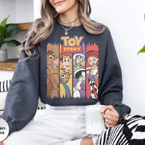 K&ouml;nnte beinhalten: Ein dunkelgrau meliertes Sweatshirt mit einem Grafikdruck des Toy Story-Logos &uuml;ber vier vertikalen Feldern. Die Felder zeigen Illustrationen von Bullseye, Woody, Buzz Lightyear und Jessie. Das Sweatshirt hat lange &Auml;rmel und einen Rundhalsausschnitt.