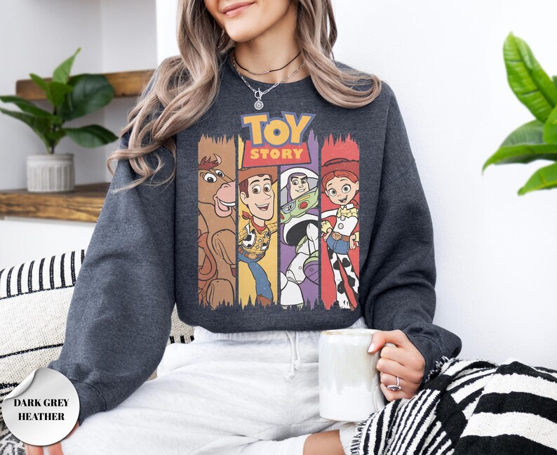 K&ouml;nnte beinhalten: Ein dunkelgrau meliertes Sweatshirt mit einem Grafikdruck des Toy Story-Logos &uuml;ber vier vertikalen Feldern. Die Felder zeigen Illustrationen von Bullseye, Woody, Buzz Lightyear und Jessie. Das Sweatshirt hat lange &Auml;rmel und einen Rundhalsausschnitt.