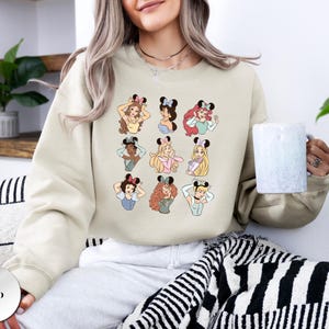 May include: Sand-colored sweatshirt featuring cartoon illustrations of various princesses wearing mouse ears. The design includes a grid of nine princess portraits, each with unique hairstyles and outfits. The sweatshirt is a neutral color.