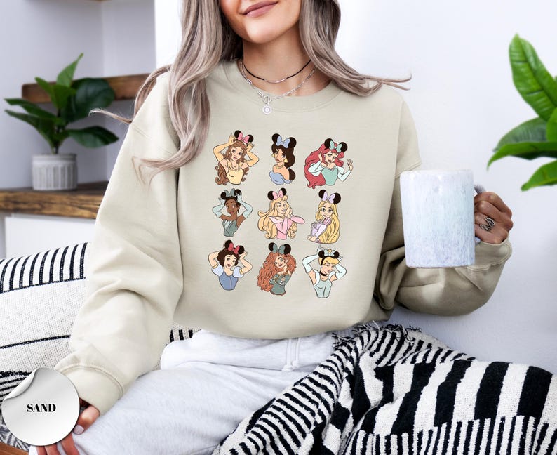 May include: Sand-colored sweatshirt featuring cartoon illustrations of various princesses wearing mouse ears. The design includes a grid of nine princess portraits, each with unique hairstyles and outfits. The sweatshirt is a neutral color.
