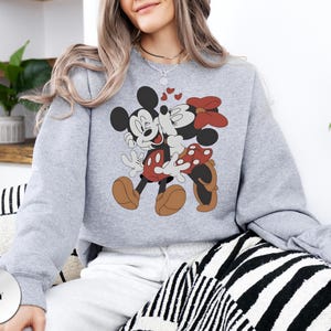 May include: A light grey sweatshirt featuring a graphic of Mickey Mouse and Minnie Mouse embracing, with hearts floating above them. Mickey is wearing red shorts and brown shoes, while Minnie wears a red polka-dot bow and shoes. The sweatshirt has a crew neck.