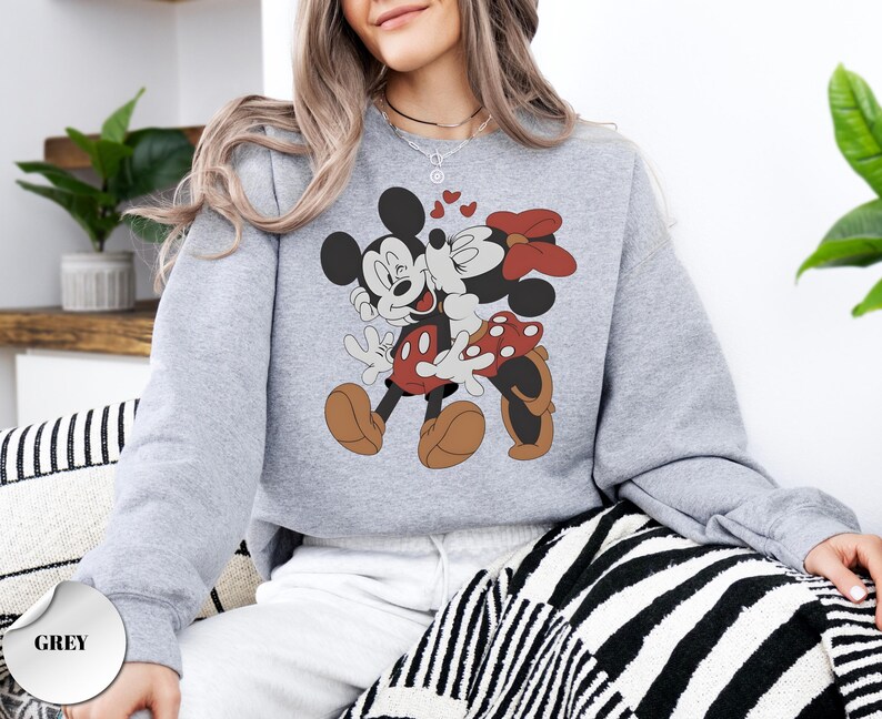May include: A light grey sweatshirt featuring a graphic of Mickey Mouse and Minnie Mouse embracing, with hearts floating above them. Mickey is wearing red shorts and brown shoes, while Minnie wears a red polka-dot bow and shoes. The sweatshirt has a crew neck.