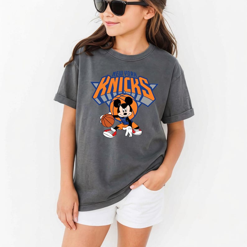 Puede incluir: Una camiseta de manga corta gris carb&oacute;n con un gr&aacute;fico de Mickey Mouse driblando un bal&oacute;n de baloncesto. Sobre el personaje, el texto NEW YORK KNICKS aparece en letras negritas de color naranja y azul. El dise&ntilde;o incluye un motivo de baloncesto.