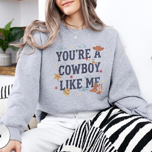 May include: A grey crewneck sweatshirt featuring the dark blue text "YOU'RE A COWBOY LIKE ME" centered on the chest. The design includes small graphic illustrations of a brown cowboy hat, a rocket ship, a yellow star, a ball, and brown cowboy boots.