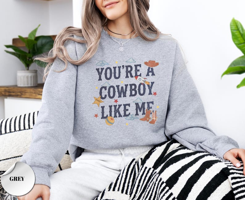 May include: A grey crewneck sweatshirt featuring the dark blue text "YOU'RE A COWBOY LIKE ME" centered on the chest. The design includes small graphic illustrations of a brown cowboy hat, a rocket ship, a yellow star, a ball, and brown cowboy boots.