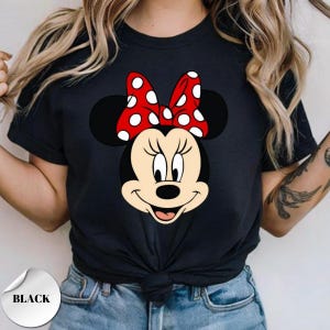 Puede incluir: Camiseta negra con un gr&aacute;fico de Minnie Mouse. El gr&aacute;fico muestra la cara de Minnie, un lazo rojo con lunares blancos. La camiseta tiene cuello redondo y mangas cortas. La palabra "BLACK" est&aacute; impresa en una pegatina blanca.