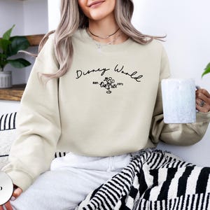 以下が含まれることがあります： 砂色のスウェットシャツ。筆記体の「Disney World」の文字、ミッキーマウスのグラフィック、そして「1971」の年号がプリントされています。クルーネックのスタイルで、柔らかい素材で作られています。