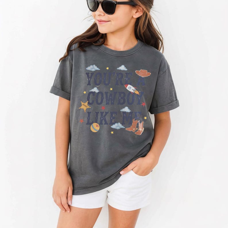 May include: A charcoal gray short-sleeve t-shirt featuring the text "YOU'RE A COWBOY LIKE ME" in dark blue letters. The graphic includes illustrations of a brown cowboy hat, a rocket ship, a yellow sheriff star, a ball, and brown cowboy boots.