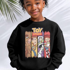 K&ouml;nnte beinhalten: Ein schwarzes Sweatshirt mit Rundhalsausschnitt und einem Grafikdruck der Toy Story-Figuren Bullseye, Woody, Buzz Lightyear und Jessie. Das Design zeigt das Toy Story-Logo in gelber und roter Schrift &uuml;ber den vier vertikalen Charakterfeldern.