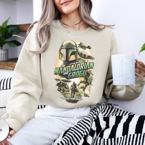 Puede incluir: Una sudadera de cuello redondo color arena con un estampado gr&aacute;fico de The Mandalorian y Grogu. El dise&ntilde;o incluye el texto "The Mandalorian and Grogu" y "This is the way" en verde y negro, con ilustraciones de los personajes y una nave espacial.