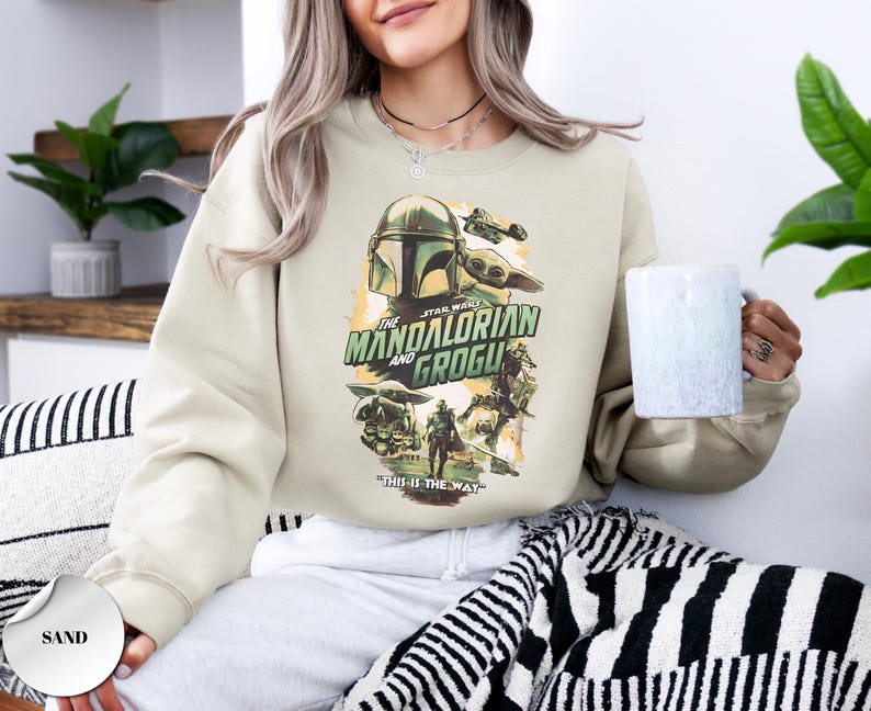 Puede incluir: Una sudadera de cuello redondo color arena con un estampado gr&aacute;fico de The Mandalorian y Grogu. El dise&ntilde;o incluye el texto "The Mandalorian and Grogu" y "This is the way" en verde y negro, con ilustraciones de los personajes y una nave espacial.