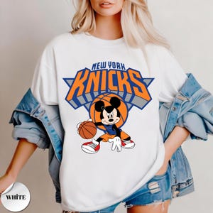 Puede incluir: Una camiseta blanca con un gr&aacute;fico de Mickey Mouse botando un bal&oacute;n de baloncesto. Sobre el personaje, el texto NEW YORK KNICKS aparece en letras grandes de color naranja y azul. El dise&ntilde;o incluye un tri&aacute;ngulo azul y un bal&oacute;n naranja.