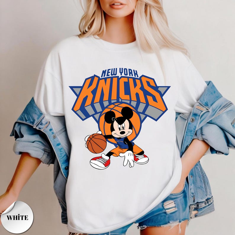 Puede incluir: Una camiseta blanca con un gr&aacute;fico de Mickey Mouse botando un bal&oacute;n de baloncesto. Sobre el personaje, el texto NEW YORK KNICKS aparece en letras grandes de color naranja y azul. El dise&ntilde;o incluye un tri&aacute;ngulo azul y un bal&oacute;n naranja.