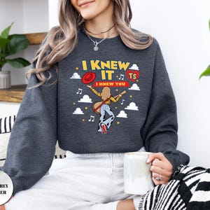 May include: A dark grey heather crewneck sweatshirt featuring a graphic print with the text I KNEW IT and I KNEW YOU. The design includes a person playing a guitar, a red hat, a red heart with TS, musical notes, stars, and white clouds.