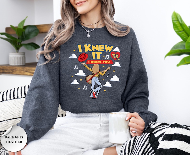May include: A dark grey heather crewneck sweatshirt featuring a graphic print with the text I KNEW IT and I KNEW YOU. The design includes a person playing a guitar, a red hat, a red heart with TS, musical notes, stars, and white clouds.