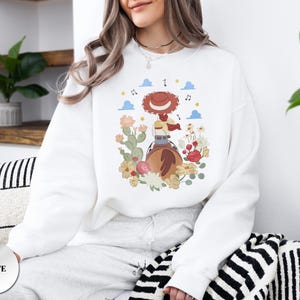 K&ouml;nnte beinhalten: Ein wei&szlig;es Sweatshirt mit Rundhalsausschnitt und einem Grafikdruck einer Cowgirl mit rotem Hut auf einem braunen Pferd. Die Illustration zeigt bunte Blumen, gr&uuml;ne Kakteen, blaue Wolken, gelbe Sterne und Noten, die die zentrale Figur umgeben.