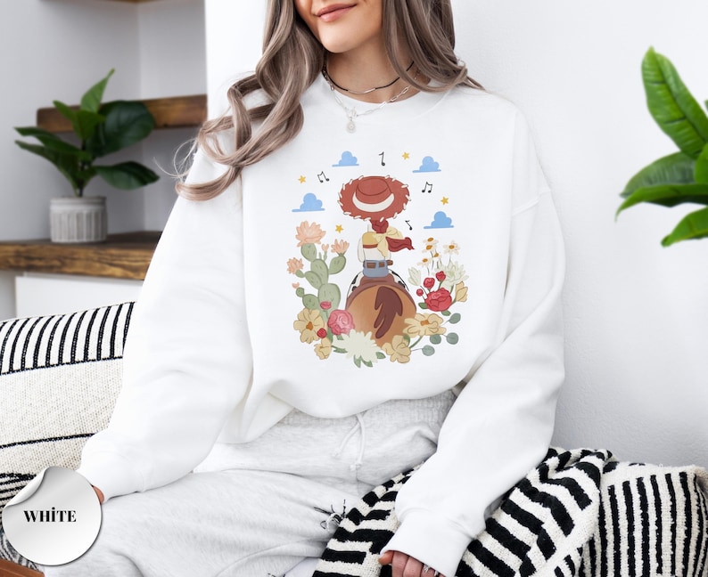 K&ouml;nnte beinhalten: Ein wei&szlig;es Sweatshirt mit Rundhalsausschnitt und einem Grafikdruck einer Cowgirl mit rotem Hut auf einem braunen Pferd. Die Illustration zeigt bunte Blumen, gr&uuml;ne Kakteen, blaue Wolken, gelbe Sterne und Noten, die die zentrale Figur umgeben.