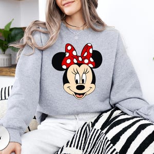 May include: A light grey sweatshirt with a Minnie Mouse graphic. The graphic shows Minnie's face, with black ears, a red bow with white spots, and a smiling expression. The word "GREY" is printed on a grey circle in the bottom left corner.