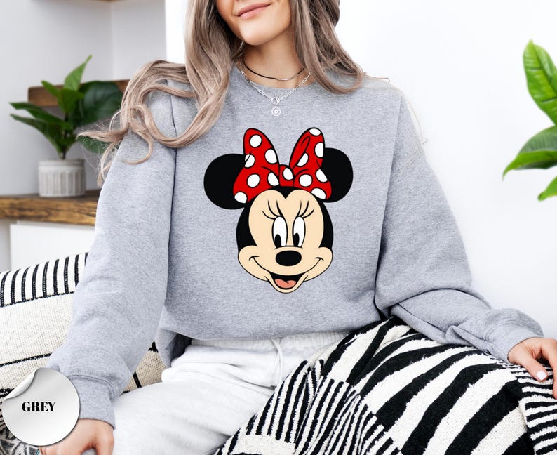 May include: A light grey sweatshirt with a Minnie Mouse graphic. The graphic shows Minnie's face, with black ears, a red bow with white spots, and a smiling expression. The word "GREY" is printed on a grey circle in the bottom left corner.
