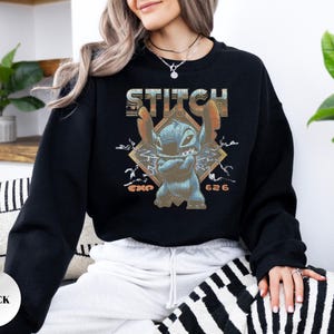 May include: A black long-sleeve crewneck sweatshirt featuring a graphic print of the character Stitch with arms crossed. The design includes the text STITCH in a distressed font, lightning bolt accents, and the text EXP 626 in orange lettering.