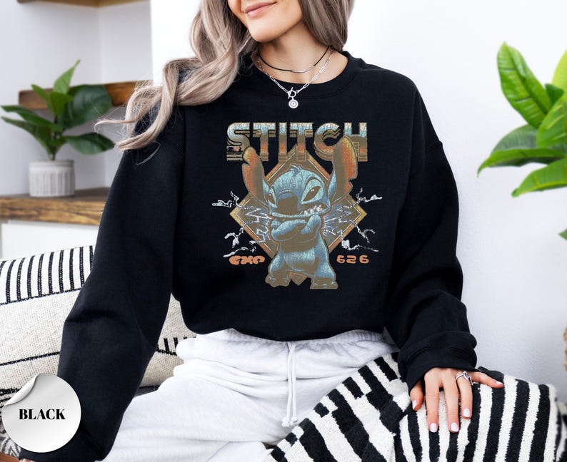 May include: A black long-sleeve crewneck sweatshirt featuring a graphic print of the character Stitch with arms crossed. The design includes the text STITCH in a distressed font, lightning bolt accents, and the text EXP 626 in orange lettering.