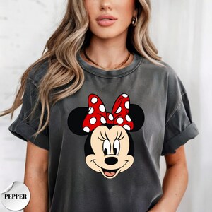 Op de afbeelding: Donkergrijs t-shirt met een Minnie Mouse-afbeelding. De afbeelding toont Minnie's gezicht met een rode strik met witte stippen. Het woord "PEPPER" staat linksonder op het shirt.