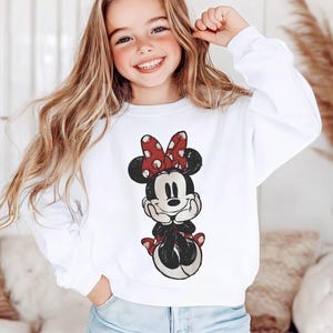 May include: White sweatshirt featuring a Minnie Mouse graphic. The graphic depicts Minnie Mouse in a seated pose, with a red bow with white polka dots. The sweatshirt is a classic crew neck style.