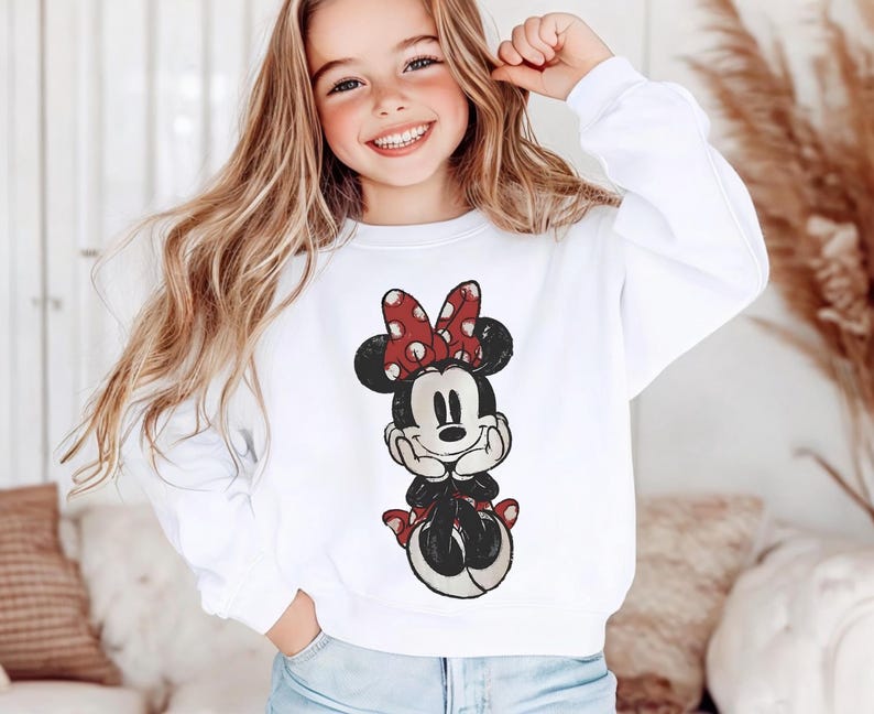 May include: White sweatshirt featuring a Minnie Mouse graphic. The graphic depicts Minnie Mouse in a seated pose, with a red bow with white polka dots. The sweatshirt is a classic crew neck style.