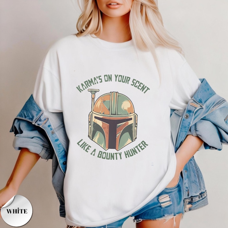 May include: A white t-shirt featuring a graphic of a stylized helmet in shades of green, orange, and brown. The text "KARMA'S ON YOUR SCENT" is printed in green above the helmet, and "LIKE A BOUNTY HUNTER" is printed in green below the helmet.