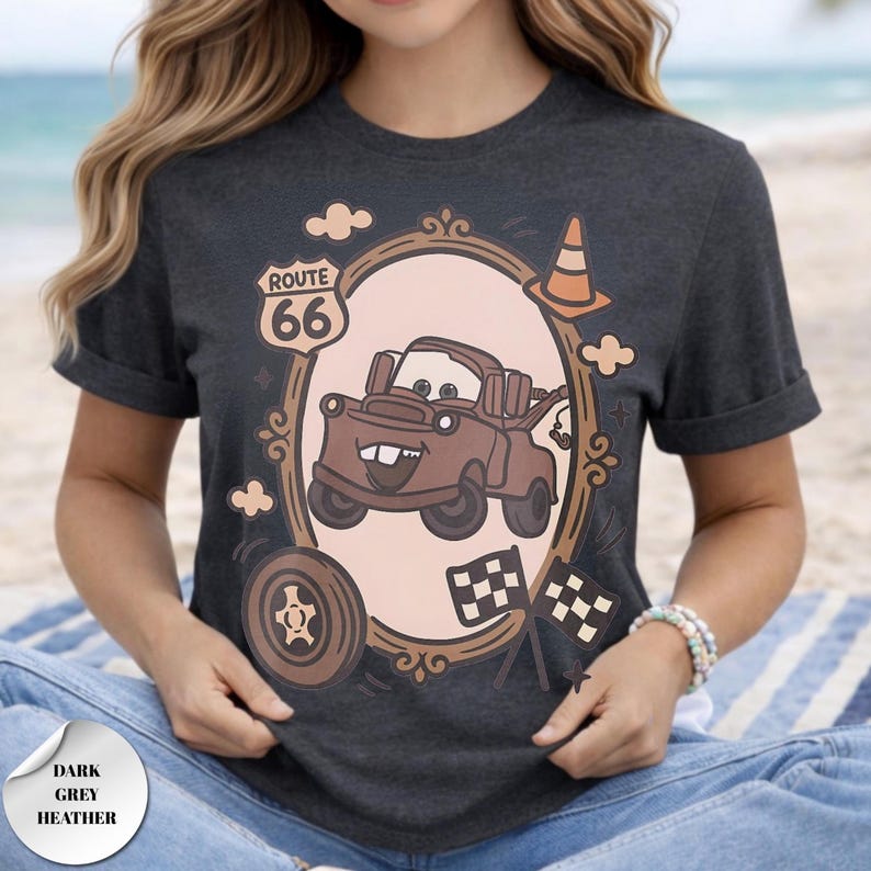 May include: A dark grey heather t-shirt featuring a graphic of a brown tow truck character inside an oval frame. The design includes a Route 66 sign, a traffic cone, a tire, and two checkered flags. The text Route 66 is printed on the sign.
