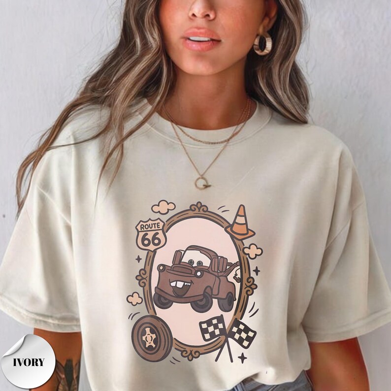 May include: An ivory short-sleeve t-shirt featuring a graphic print of a brown tow truck character inside an oval frame. The design includes a Route 66 sign, a traffic cone, a tire, and two checkered flags. The shirt has a crew neckline.