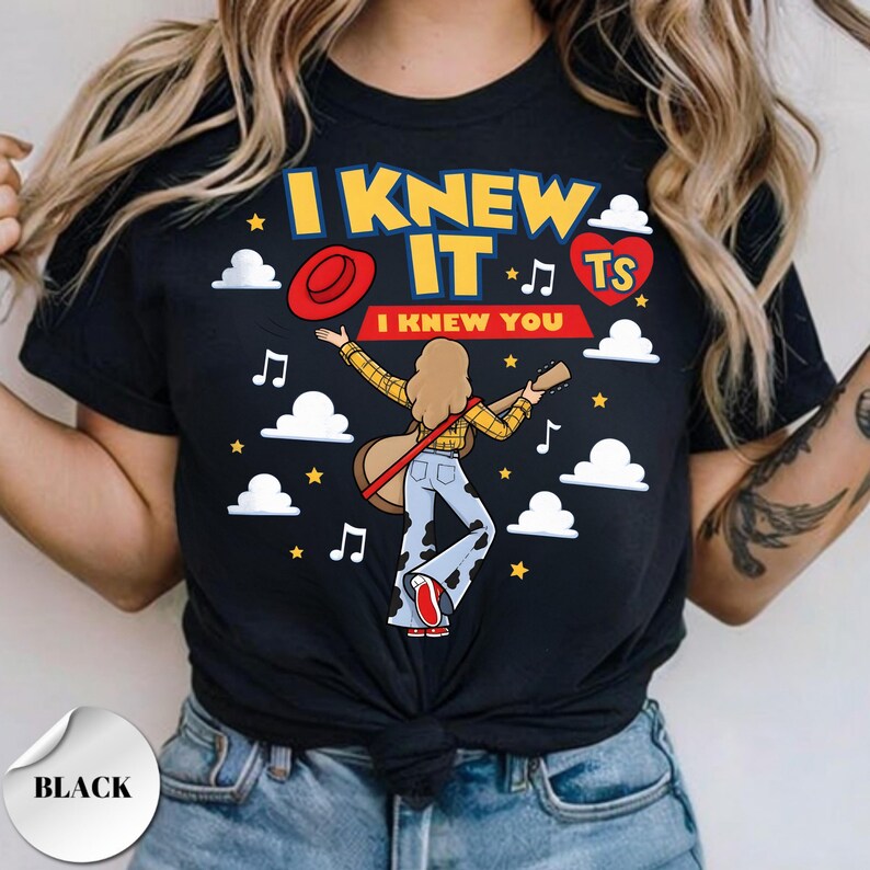 May include: A black t-shirt featuring a graphic of a person with long hair wearing a yellow plaid shirt and cow-print bell-bottom pants, holding a guitar. The text reads I KNEW IT and I KNEW YOU, accompanied by a red hat, heart, stars, and clouds.