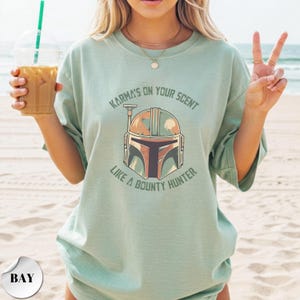 May include: A light sage green short-sleeve t-shirt featuring a graphic of a helmet in the center. The text above the helmet reads "KARMA'S ON YOUR SCENT" and the text below reads "LIKE A BOUNTY HUNTER" in a dark green, distressed font.