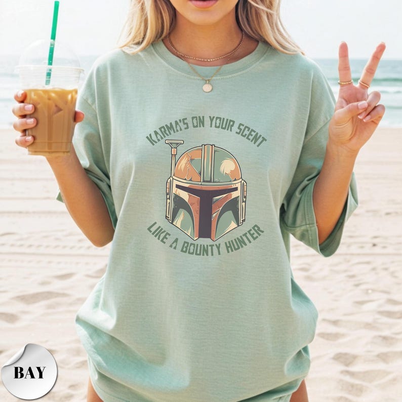 May include: A light sage green short-sleeve t-shirt featuring a graphic of a helmet in the center. The text above the helmet reads "KARMA'S ON YOUR SCENT" and the text below reads "LIKE A BOUNTY HUNTER" in a dark green, distressed font.