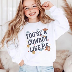 May include: A white long-sleeve crewneck sweatshirt featuring the dark blue text "YOU'RE A COWBOY LIKE ME" centered on the chest. The design includes small colorful illustrations of a cowboy hat, a rocket, a sheriff star, boots, and a ball.