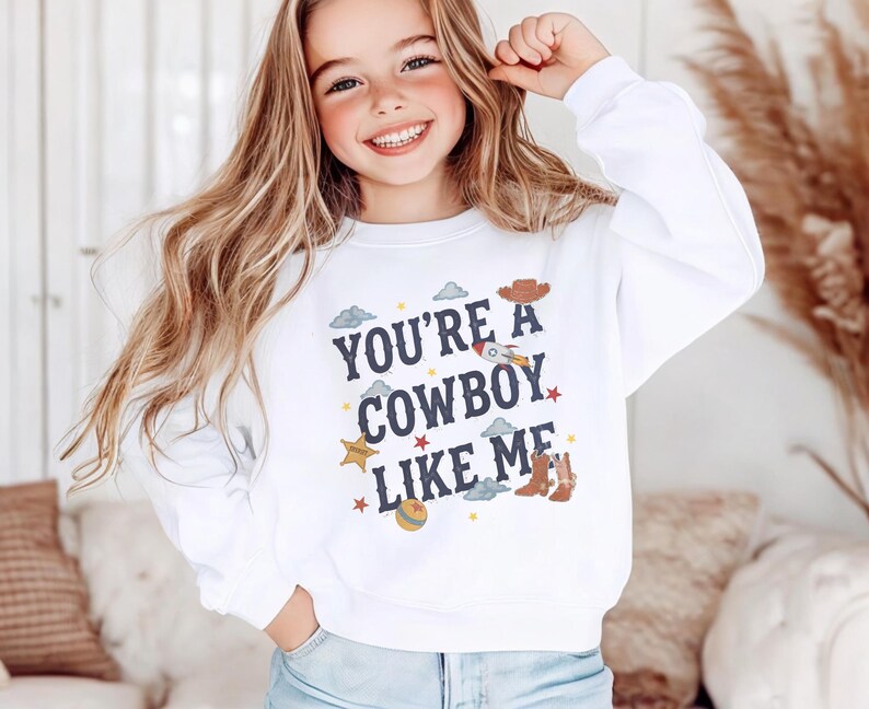 May include: A white long-sleeve crewneck sweatshirt featuring the dark blue text "YOU'RE A COWBOY LIKE ME" centered on the chest. The design includes small colorful illustrations of a cowboy hat, a rocket, a sheriff star, boots, and a ball.