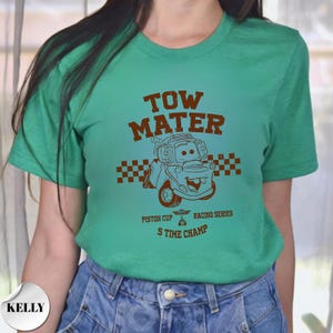 May include: A bright green short-sleeve t-shirt featuring a brown graphic of the character Tow Mater from the movie Cars. The design includes a checkered flag, the text TOW MATER, PISTON CUP RACING SERIES, and 5 TIME CHAMP in brown ink.