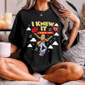 Puede incluir: Una camiseta negra con un estampado gr&aacute;fico con el texto I KNEW IT y I KNEW YOU. El dise&ntilde;o incluye una persona con cabello largo tocando la guitarra, un sombrero rojo, un coraz&oacute;n rojo con TS, nubes blancas, estrellas amarillas y notas musicales.