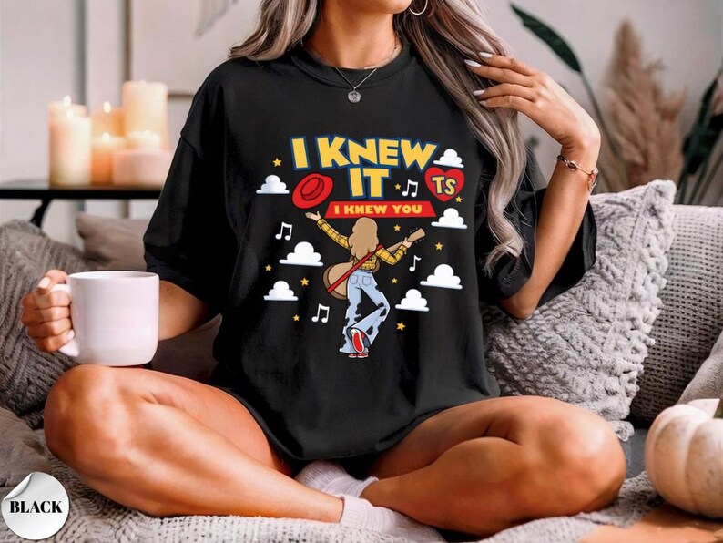 Puede incluir: Una camiseta negra con un estampado gr&aacute;fico con el texto I KNEW IT y I KNEW YOU. El dise&ntilde;o incluye una persona con cabello largo tocando la guitarra, un sombrero rojo, un coraz&oacute;n rojo con TS, nubes blancas, estrellas amarillas y notas musicales.